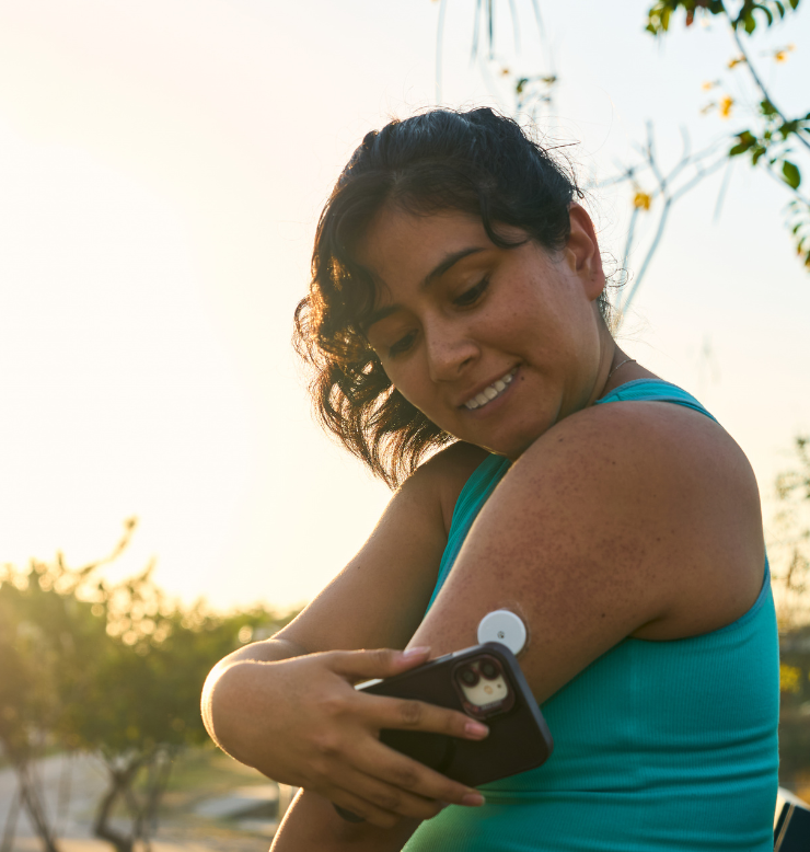 Photo shows a woman in a teal tank-top, using her phone to check the continuous glucose monitor on her left arm. 